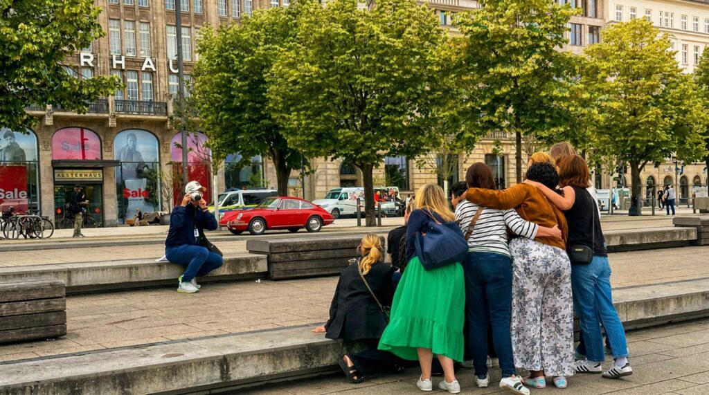 Eine Gruppe Menschen wird fotografiert von einem Tour-Guide mit einer Polaroid-Kamera. Das Foto entstand bei einer Stadtrallye-Station, wo eine Aufgabe zu lösen war. Die Stadtrallye in Hamburg wurde von den Rosinenfischern organisiert.Das Foto wurde während des Team Events bei dieser Hamburg Stadtrallye bzw. Schnitzeljagd auf dem Jungfernstieg aufgenommen.